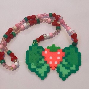 May include: A necklace featuring a central pendant made of fused plastic beads in the shape of a red strawberry with green leaves. The necklace strand consists of a repeating pattern of red, pink, green, and clear beads with small red heart beads.