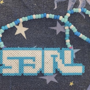 Puede incluir: Un collar de cuentas con las letras "SNL" en un diseño pixelado. Las letras son de color azul claro y blanco, con una cadena de cuentas a juego en tonos de azul y blanco. El fondo es una tela azul oscuro con patrones de estrellas y luna.