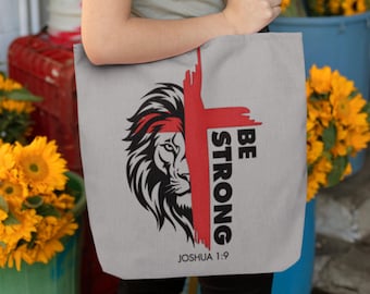May include: A gray canvas tote bag with a black and white lion graphic and a red cross. The text "BE STRONG" is written vertically in black on the cross. The text "JOSHUA 1:9" is written in black at the bottom of the bag.