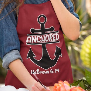 Op de afbeelding: Een rode schort met een zwart anker en de woorden "Anchored" en "Hebreeën 6:19" in wit.