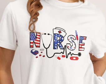 Puede incluir: Camiseta blanca con la palabra "NURSE" en un diseño patriótico. Las letras están decoradas con la bandera americana y estrellas. Un estetoscopio, pastillas y una jeringa también aparecen en el diseño.