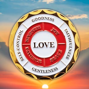 May include: A gold-colored round coin with the word "LOVE" in the center, surrounded by a red and white design. The coin features words like "GOODNESS," "JOY," and "FAITHFULNESS" around the edges. The background shows a sunset.