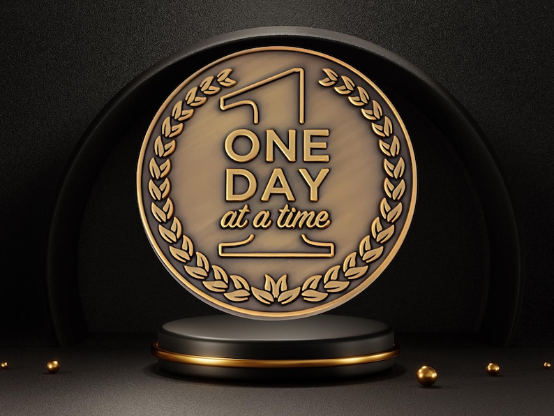 May include: A bronze-colored medallion with the words "ONE DAY at a time" in gold lettering, surrounded by a laurel wreath. The medallion rests on a black pedestal with a gold trim, set against a dark background.