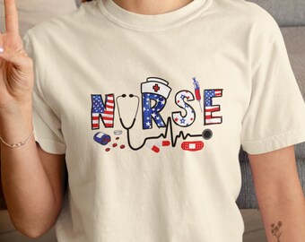 Puede incluir: Camiseta color crema con la palabra "NURSE" en letras rojas, blancas y azules. El diseño incluye un estetoscopio, un gorro de enfermera, una jeringa, pastillas y una tirita, todo en colores patrióticos.