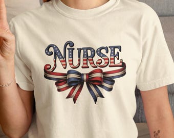 Puede incluir: Camiseta color crema con la palabra "NURSE" en un diseño patriótico con colores rojo, blanco y azul. La palabra está arqueada sobre un lazo de cinta con la misma combinación de colores. Un diseño para profesionales de la salud.
