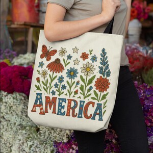 Könnte beinhalten: Cremefarbene Tragetasche mit floralem Design und dem Wort "AMERICA" in Rot, Weiß und Blau. Das Design umfasst einen Schmetterling, Gänseblümchen und Sterne. Die Tasche hat schwarze Griffe und wird von einer Person gehalten. Das florale Design umfasst Gänseblümchen, Sterne und andere Blumen.