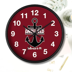 Könnte beinhalten: Eine rote runde Wanduhr mit einem schwarzen Ankerdesign und den Worten "Anchored" und "Hebrews 6:19". Die Uhr hat schwarze Zeiger und weiße Zahlen.