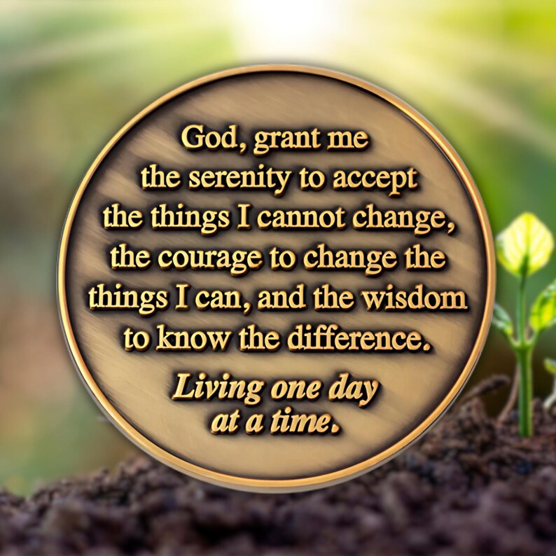 May include: A round, bronze-colored coin with a quote in black text: "God, grant me the serenity to accept the things I cannot change... Living one day at a time." The coin is set against a blurred green and brown background.