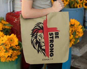 May include: Beige canvas tote bag with a black and white lion graphic and a red cross. The text "BE STRONG" is printed vertically in black and "JOSHUA 1:9" is printed in black at the bottom.
