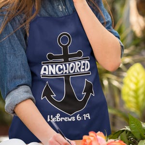 Op de afbeelding: Een marineblauwe schort met een wit ankerontwerp en de tekst "Anchored Hebrews 6:19".