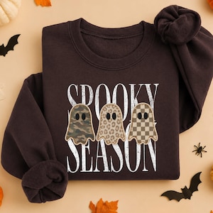 May include: A dark chocolate brown crewneck sweatshirt featuring the white text SPOOKY SEASON and three ghost illustrations. The ghosts have distinct patterns: one in camouflage, one in leopard print, and one in a tan and brown checkerboard design.