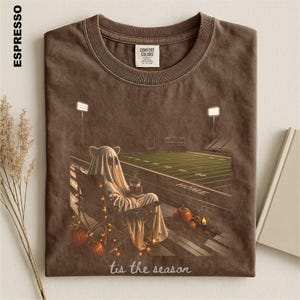 May include: A brown t-shirt featuring a graphic of a ghost sitting in a camping chair on a football field at night. The ghost holds a drink, surrounded by pumpkins and string lights. The text 'tis the season' is printed at the bottom in white.