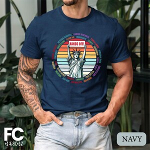 May include: A navy blue t-shirt with a vintage-style graphic of the Statue of Liberty holding a sign that says "Hands Off!" The graphic is surrounded by a circle of text that includes phrases like "Clean Air", "Medicare", "Our Jobs", and "Fair Elections".