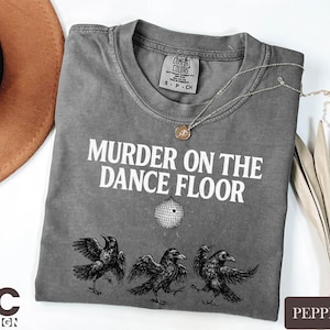 May include: Pepper-colored t-shirt with the text "MURDER ON THE DANCE FLOOR" and a disco ball graphic. Below the text are three crow illustrations. A gold necklace is draped across the shirt. A brown hat is in the upper left corner.