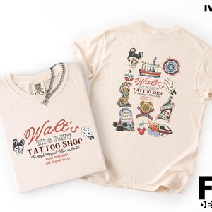 May include: Ivory-colored t-shirts with a tattoo shop theme. One shirt displays the text "Walt's Ink & Paint Tattoo Shop" with a skull and Mickey Mouse hand graphic. The other shirt features various tattoo-style illustrations.
