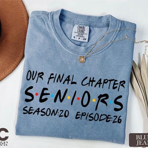 May include: A blue jean colored t-shirt with the text "OUR FINAL CHAPTER SENIORS SEASON:20 EPISODE:26" in black. The word "SENIORS" has colored dots in place of the letters "O" and "I". A gold necklace is draped across the shirt.