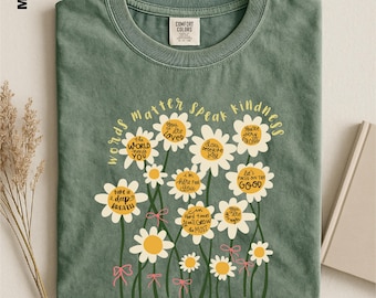 May include: A moss green t-shirt featuring a graphic of white daisies with yellow centers. The text "words matter SPEAK kindness" is arched above the flowers, and each daisy center contains phrases like "You are LOVED" and "THE WORLD NEEDS YOU."