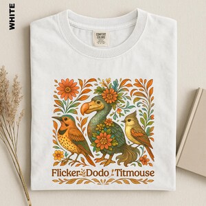 May include: A white t-shirt featuring a graphic illustration of a flicker, a dodo, and a titmouse surrounded by orange and green floral elements. The text "Flicker Dodo Titmouse" is printed in brown at the bottom of the graphic design.