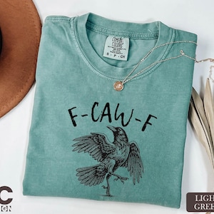 A turquoise t-shirt with a graphic of a bird and the text "F-CAW-F" printed on it, along with a necklace and a brown hat.