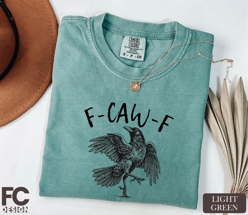 A turquoise t-shirt with a graphic of a bird and the text "F-CAW-F" printed on it, along with a necklace and a brown hat.