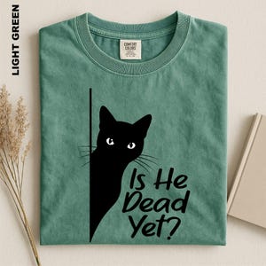 May include: A light green crew neck t-shirt featuring a black graphic of a cat peeking from behind a vertical line. The text "Is He Dead Yet?" is printed in black next to the cat. The shirt has a Comfort Colors brand label at the neckline.
