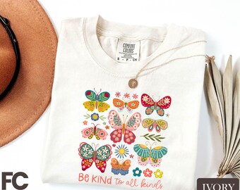 May include: Ivory-colored t-shirt with a colorful butterfly graphic and the text "Be Kind to all kinds." The butterflies are in various colors and designs. A gold necklace is draped on the shirt.