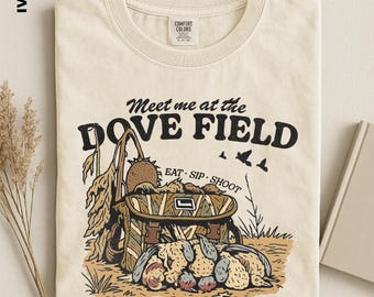 May include: An ivory t-shirt featuring a graphic print with the text "Meet me at the DOVE FIELD" and "EAT SIP SHOOT". The illustration depicts a camouflage bag, sunflowers, and birds in a field setting. The shirt has a crew neck and a label.