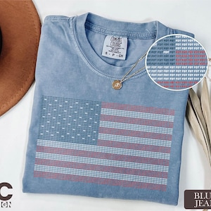 May include: Blue jean t-shirt with an American flag design. The flag is made up of the words "FOT" in blue and red. The shirt has a crew neck and short sleeves. A gold necklace with a pendant is on the shirt.
