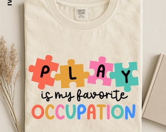 May include: An ivory t-shirt featuring the text "PLAY is my favorite OCCUPATION". The word "PLAY" is written in black letters inside four colorful puzzle pieces in red, yellow, pink, and teal. The word "OCCUPATION" appears below in multicolored letters.