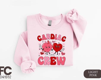 Puede incluir: Sudadera rosa claro con un gráfico de dos corazones de dibujos animados tomados de la mano. El texto "Cardiac Crew" está impreso en letras rosa y rojo.