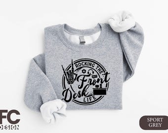 May include: Grey sweatshirt with the slogan "Rocking the Front Desk Life" in a black circular design. The design includes a skeleton hand making a rock and roll gesture. The sweatshirt has white cuffs.
