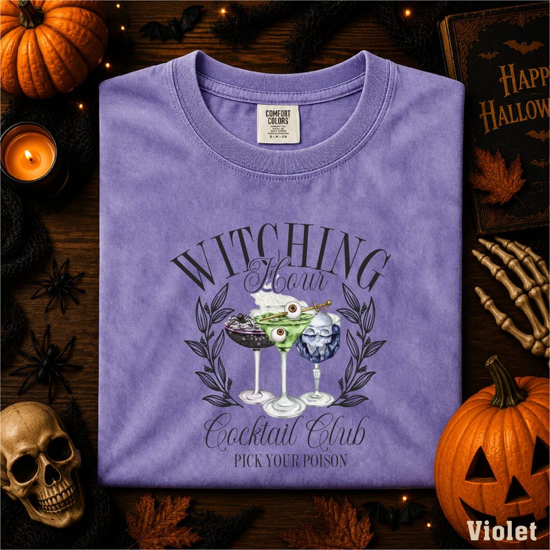 a purple t-shirt with a graphic design of cocktail glasses and a witch's hat, surrounded by Halloween decorations such as a pumpkin, bats, and a skeleton.