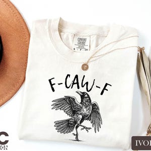 a white t-shirt with a graphic design of a bird, specifically a crow, on it. The t-shirt is placed on a white background, along with a brown hat and some leaves.