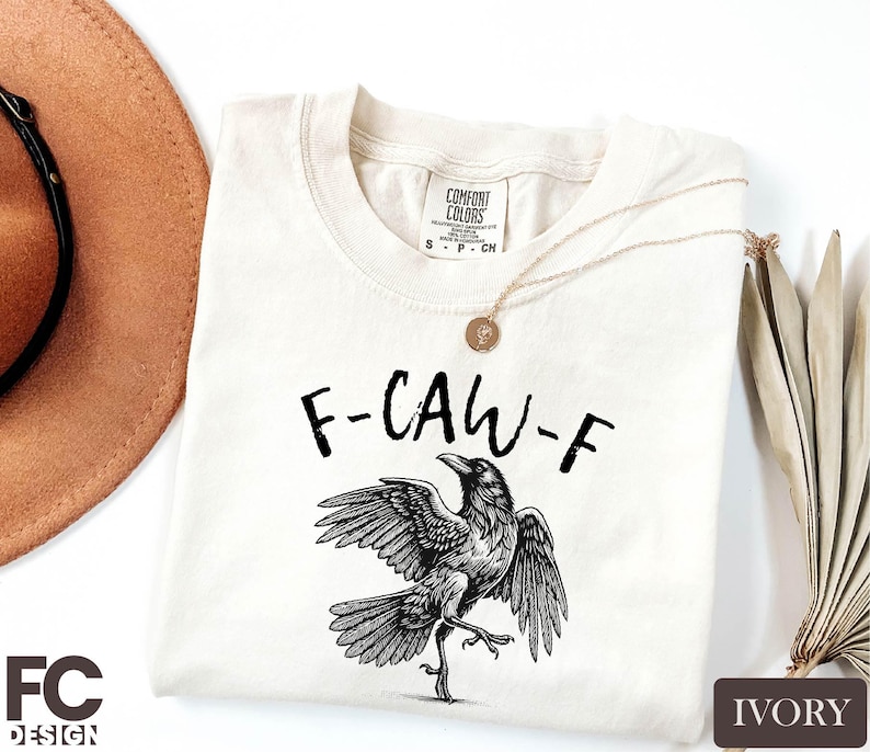 a white t-shirt with a graphic design of a bird, specifically a crow, on it. The t-shirt is placed on a white background, along with a brown hat and some leaves.