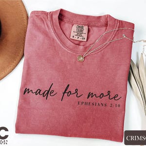 a pink t-shirt with the text "made for more" printed on it, along with a small pendant necklace. The t-shirt is placed on a white surface, and there is a brown hat and some dried leaves nearby.