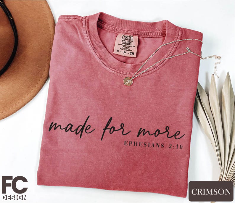 a pink t-shirt with the text "made for more" printed on it, along with a small pendant necklace. The t-shirt is placed on a white surface, and there is a brown hat and some dried leaves nearby.