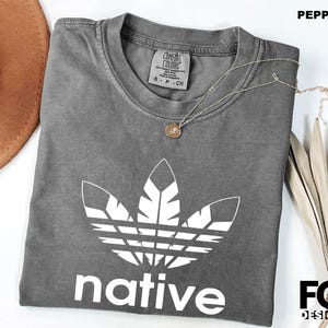 May include: A gray t-shirt with a white graphic design and the word "native" printed below. The graphic is a stylized feather design. A gold necklace with a pendant rests on the shirt. The shirt is labeled "Pepper".