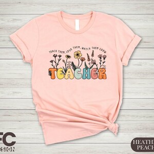 May include: Pink t-shirt with the text "Teach them, love them, watch them grow" and the word "Teacher" in colorful letters with flowers around the text.