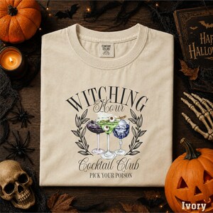 a white t-shirt with a graphic design of a cocktail glass and text that says "WITCHING" and "Cocktail Club" along with the phrase "Pick your poison". The t-shirt is laid out on a wooden surface, surrounded by Halloween decorations such as a pumpkin, bats, and a skeleton.