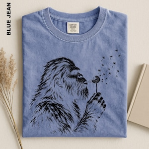 A blue t-shirt with a black graphic design depicting a furry creature blowing a dandelion seed into the air.