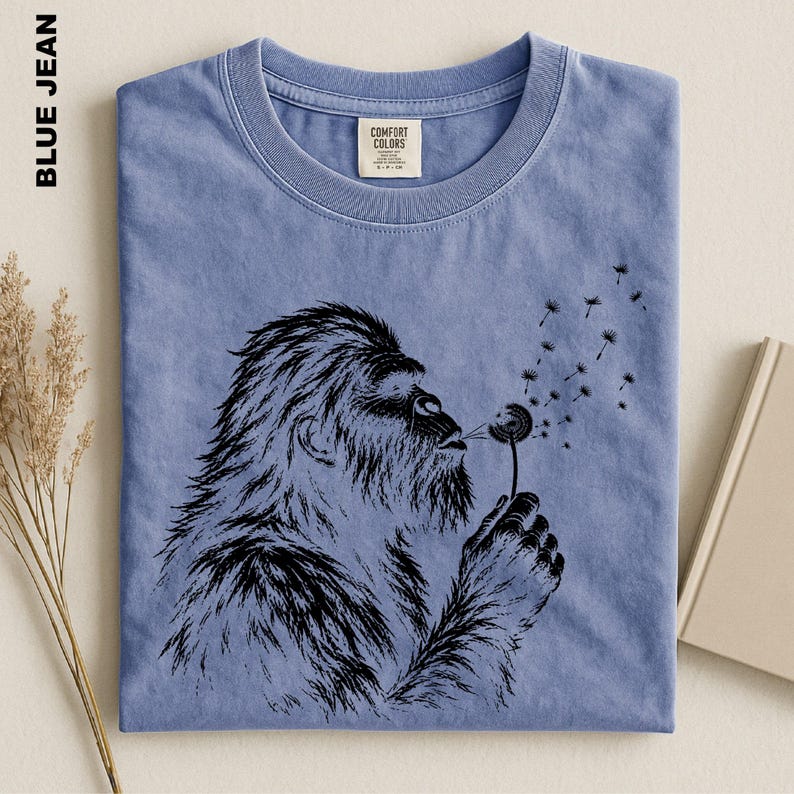 A blue t-shirt with a black graphic design depicting a furry creature blowing a dandelion seed into the air.