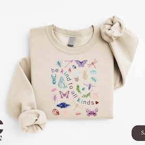 May include: Sand-colored sweatshirt with a crew neck and long sleeves. The front features a colorful illustration of various insects and the phrase "be kind to all kinds". The design includes butterflies, dragonflies, and beetles.