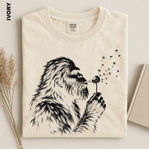 a t-shirt with a black and white illustration of a furry animal blowing a dandelion, with the dandelion seeds floating around it.