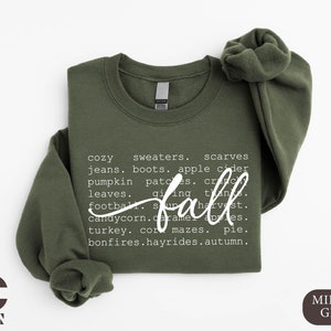 May include: A green sweatshirt with white text that reads "cozy sweaters, scarves, jeans, boots, apple cider, pumpkin patches, crunching leaves, giving thanks, football, fall, candy corn, caramel apples, turkey, corn mazes, pie, bonfires, hayrides, autumn."