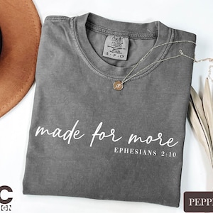 a gray t-shirt with the text "made for more" printed on it, along with the Bible verse "Ephesians 2:10" below it. The t-shirt is laid out on a white surface, accompanied by a brown hat, a necklace, and some dried leaves.