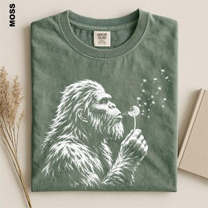 A green t-shirt with a white illustration of a gorilla blowing a dandelion, accompanied by a book and some dried flowers.