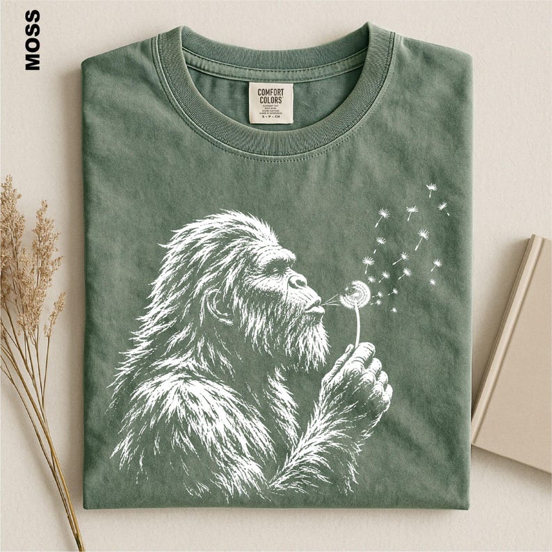 A green t-shirt with a white illustration of a gorilla blowing a dandelion, accompanied by a book and some dried flowers.
