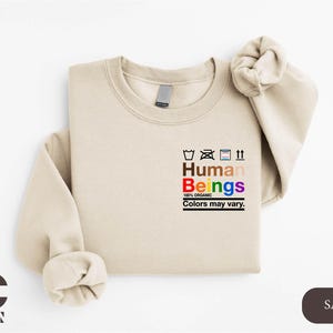 May include: A sand-colored sweatshirt with the text "Human Beings 100% Organic Colors may vary." printed on the front. The text is in black and rainbow colors.