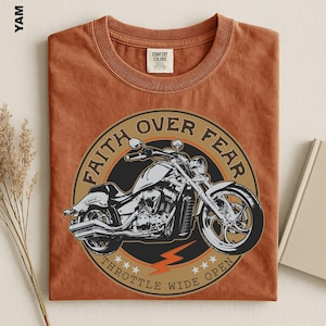 May include: A rust-colored t-shirt featuring a circular graphic of a motorcycle. The text reads FAITH OVER FEAR and THROTTLE WIDE OPEN, accompanied by a lightning bolt and stars. The shirt has a crew neck and a Comfort Colors brand label inside.