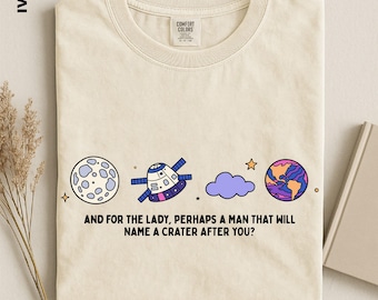 Puede incluir: Una camiseta de color marfil con un gráfico de la luna, una nave espacial, una nube morada y un planeta colorido. Debajo del gráfico, el texto dice: AND FOR THE LADY, PERHAPS A MAN THAT WILL NAME A CRATER AFTER YOU? La camiseta tiene una etiqueta Comfort Colors.